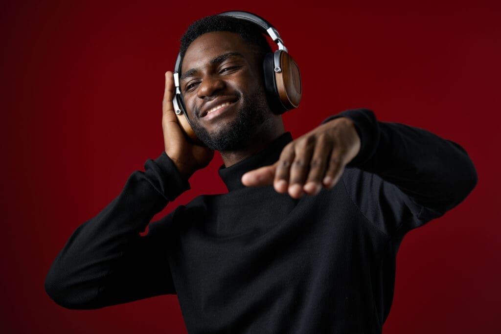 A young Black man immersed in music, wearing modern headphones, smiles joyfully against a striking red backdrop, conveying passion, style, and positive energy