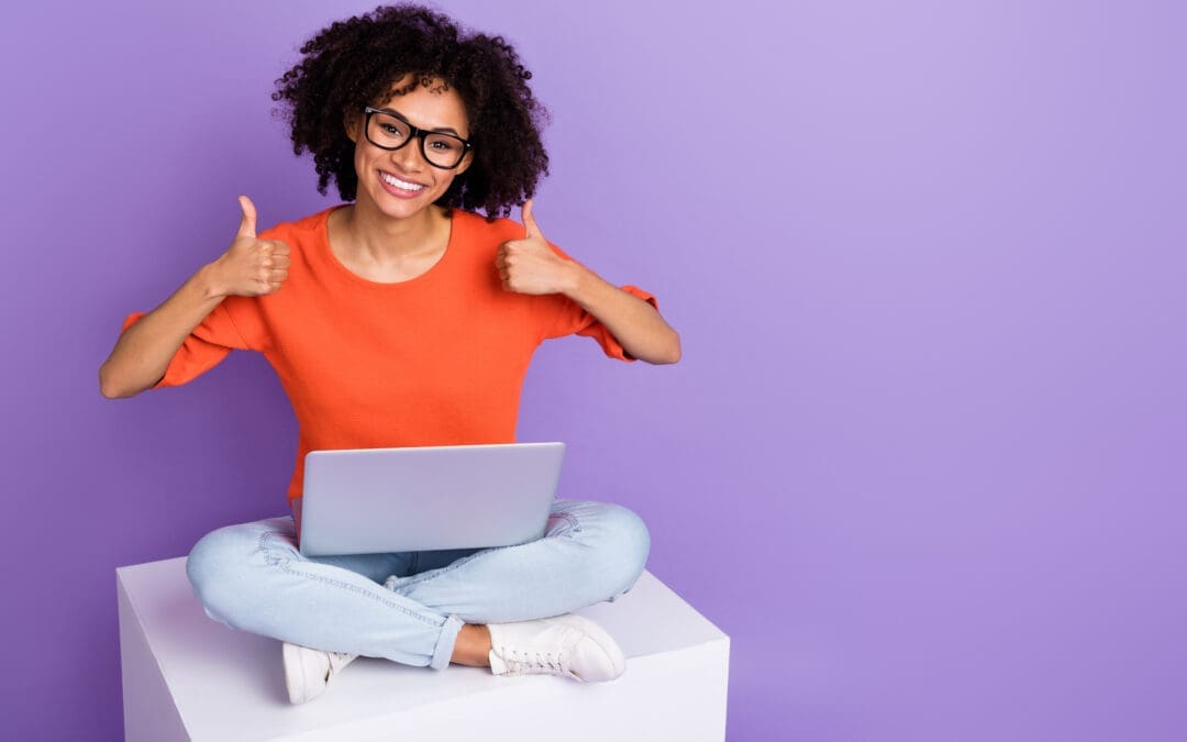 Woman with black curly hair sitting on a box with a laptop. She has both thumbs up. There is a purple background and the woman is wearing an orange shirt.